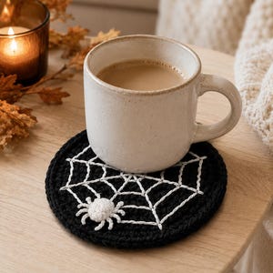 May include: A round, black crocheted coaster featuring a white embroidered spider web pattern and a small, three-dimensional white crocheted spider attached to the surface, supporting a speckled, light-coloured ceramic mug filled with coffee.