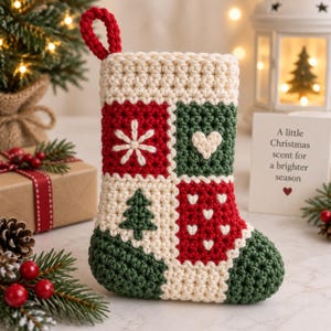 May include: A crochet Christmas stocking featuring a patchwork design in cream, red, and dark green. The stocking displays embroidered motifs including a white snowflake, a cream heart, a green pine tree, and small white hearts on a red background.