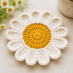 May include: A round, crocheted coaster shaped like a daisy flower. The centre is made of bright yellow yarn in a circular pattern, surrounded by twelve rounded, white yarn petals. The texture is consistent with hand-knitted cotton yarn.