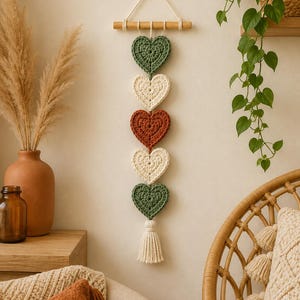 May include: A vertical wall hanging featuring five crocheted hearts in alternating colours of sage green, cream, and terracotta, suspended from a horizontal wooden dowel by white cotton cord, finished with a cream-coloured tassel at the bottom.