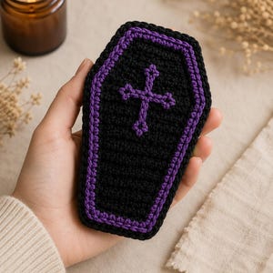May include: A small, hand-crocheted coffin-shaped item featuring a black base with a vibrant purple border and a matching purple cross stitched in the center. The piece is crafted with a textured yarn finish, showcasing a detailed, uniform stitch pattern.