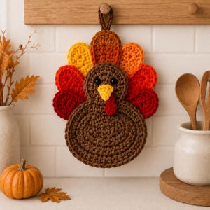May include: A crocheted turkey-shaped potholder featuring a brown body, a yellow beak, a red wattle, and black eyes. The tail feathers are arranged in a fan shape using alternating colours of yellow, orange, red, and dark red yarn.