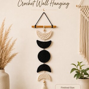 Pode incluir: Uma tapeçaria de crochê com cinco fases da lua em fio creme e preto, suspensa por um bastão de madeira com um cordão preto. O texto diz Moon Phase Crochet Wall Hanging e Finished Size: 8 in W x 18 in H, com uma borla creme na base inferior.