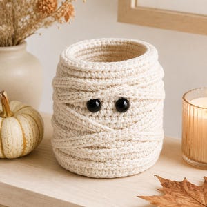 May include: A cylindrical crochet basket crafted from off-white yarn, featuring a mummy-inspired design with layered yarn strips wrapped around the exterior and two small, round, glossy black plastic eyes positioned near the top centre.