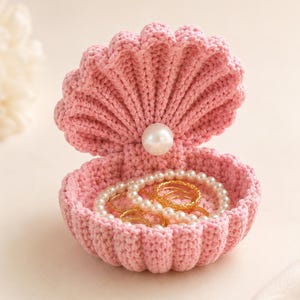 May include: A light pink crocheted seashell container with a hinged lid. A single white faux pearl is attached to the center of the upper shell. Inside the bottom shell, there is a collection of gold-toned metal rings and a white pearl bead necklace.