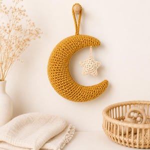 Pode incluir: Um enfeite de parede em forma de meia-lua de crochê na cor amarelo-mostarda, apresentando uma pequena estrela de crochê na cor creme suspensa no centro. A lua possui uma alça de fio amarelo-mostarda correspondente no topo para pendurar.