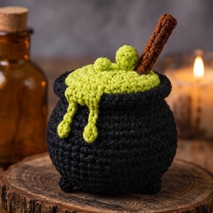 Op de afbeelding: Een zwarte gehaakte ketel met een getextureerd oppervlak en drie kleine voetjes. De bovenkant is gevuld met fel limoengroene gehaakte vloeistof die over de rand druipt, met twee kleine groene bolletjes en een bruine getextureerde stok die omhoog steekt.