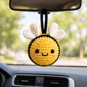 May include: A round, crocheted bee ornament hanging from a black loop. The bee has a yellow body with black stripes, white heart-shaped wings, two black bead eyes, and small pink stitched cheeks, creating a cheerful, smiling facial expression.