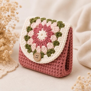 May include: A small crochet pouch featuring a square granny motif flap in cream, dusty rose, and olive green yarn. The main body of the pouch is solid dusty rose crochet, and the flap is secured with a single round, light brown wooden button.