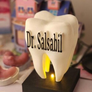 May include: A white, tooth-shaped lamp with the text "Dr. Salsabil" in black. The lamp is on a black base and glows with a warm, yellow light. The background features miniature houses and dental molds.