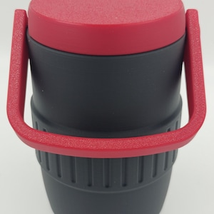 Puede incluir: Un termo de viaje aislado negro y rojo con tapa y asa rojas texturizadas. El termo tiene forma cil&iacute;ndrica con una secci&oacute;n inferior acanalada y un asa que se arquea sobre la parte superior.