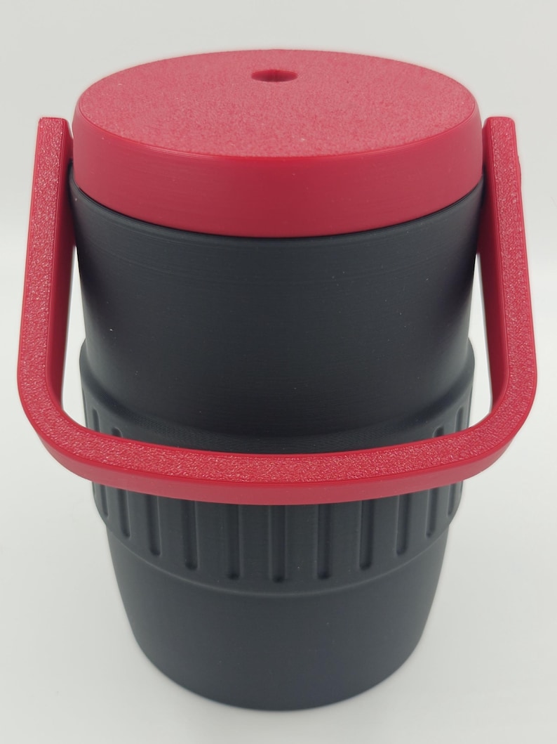 Puede incluir: Un termo de viaje aislado negro y rojo con tapa y asa rojas texturizadas. El termo tiene forma cil&iacute;ndrica con una secci&oacute;n inferior acanalada y un asa que se arquea sobre la parte superior.