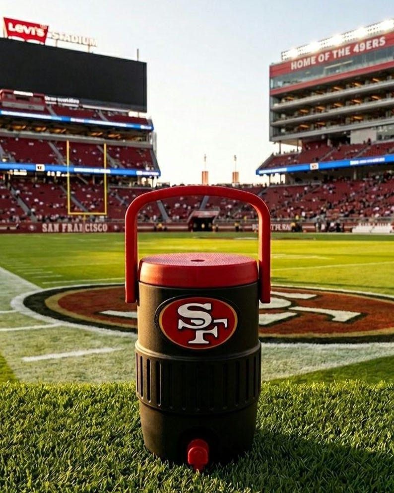 Puede incluir: Un enfriador de agua negro y rojo con el logotipo de los 49ers de San Francisco, un asa y un grifo. El enfriador est&aacute; en un campo verde con el logotipo de los 49ers y el estadio Levi's al fondo.