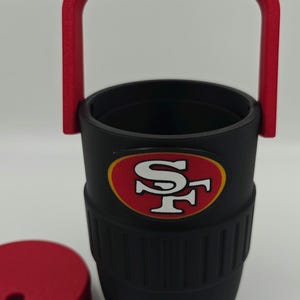 Puede incluir: Vaso t&eacute;rmico negro con asa y tapa rojas. El vaso presenta el logotipo de los 49ers de San Francisco en rojo y dorado. El asa es arqueada y la tapa es redonda. El vaso est&aacute; dise&ntilde;ado para mantener las bebidas fr&iacute;as o calientes.