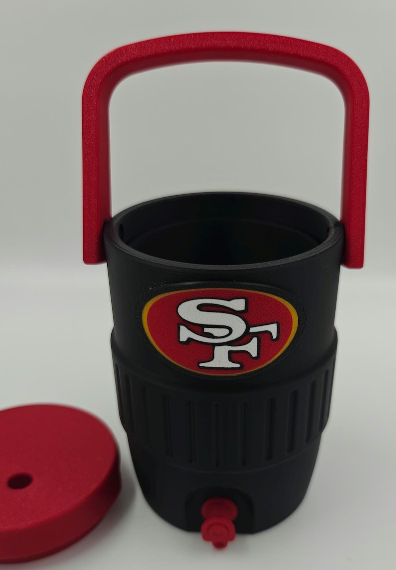 Puede incluir: Vaso t&eacute;rmico negro con asa y tapa rojas. El vaso presenta el logotipo de los 49ers de San Francisco en rojo y dorado. El asa es arqueada y la tapa es redonda. El vaso est&aacute; dise&ntilde;ado para mantener las bebidas fr&iacute;as o calientes.