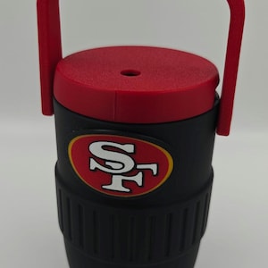 Puede incluir: Enfriador de agua aislado negro y rojo con asa. El enfriador presenta el logotipo de los 49ers de San Francisco en rojo y dorado. Tiene una tapa roja y un grifo para dispensar l&iacute;quidos.
