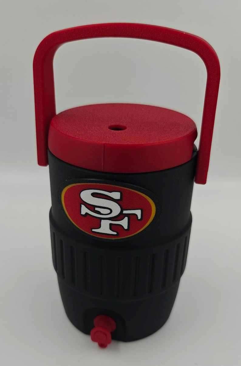 Puede incluir: Enfriador de agua aislado negro y rojo con asa. El enfriador presenta el logotipo de los 49ers de San Francisco en rojo y dorado. Tiene una tapa roja y un grifo para dispensar l&iacute;quidos.