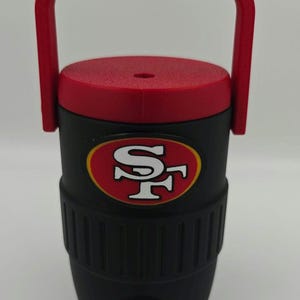 Puede incluir: Enfriador de agua aislado negro y rojo con asa. El enfriador presenta el logotipo de los 49ers de San Francisco en la parte delantera. Tiene una tapa y un asa rojas, y un grifo en la parte inferior para dispensar l&iacute;quidos.