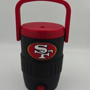 Puede incluir: Nevera de agua aislada negra y roja con asa y tapa rojas. La nevera presenta el logotipo de los 49ers de San Francisco en la parte delantera. Tiene un grifo para dispensar l&iacute;quidos.