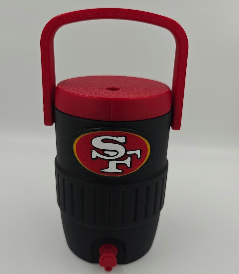 Puede incluir: Nevera de agua aislada negra y roja con asa y tapa rojas. La nevera presenta el logotipo de los 49ers de San Francisco en la parte delantera. Tiene un grifo para dispensar l&iacute;quidos.