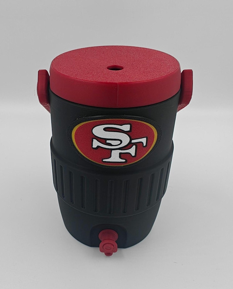 Puede incluir: Enfriador de agua aislado negro y rojo con el logotipo de los 49ers de San Francisco. El enfriador tiene una tapa y asas rojas, un cuerpo negro y un grifo. El logotipo del equipo se muestra en la parte delantera.