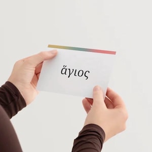 May include: A white flashcard with the Greek word "άγιος" in black lettering. The card has a colorful stripe at the top and is held by two hands against a white background.