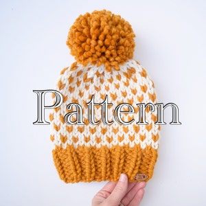 May include: A white and mustard yellow knitted beanie with a pom pom on top. The beanie has a geometric pattern and the word "Pattern" is written in large letters across the front.
