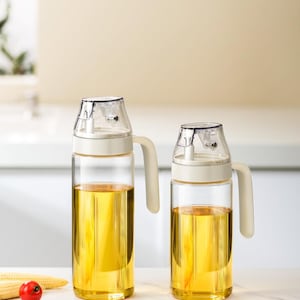 May include: Two clear glass oil dispensers with white lids and handles. The dispensers are filled with yellow oil. A small red tomato and corn on the cob are in the foreground. The dispensers are on a white surface.