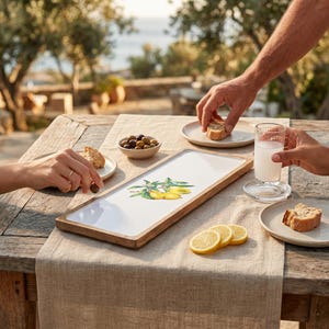May include: A rectangular wooden serving platter with a white surface featuring an olive branch illustration. The table setting includes plates of food, olives, and lemon slices. A glass of milk is also visible.