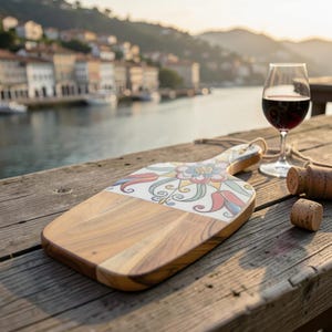 Op de afbeelding: Houten serveerplank met een decoratief bloemmotief. De plank heeft een lichtbruine houten basis met een wit keramisch gedeelte met een kleurrijk bloemenpatroon. Een glas rode wijn en kurken zijn ook zichtbaar.
