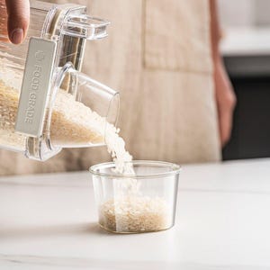 Op de afbeelding: Een doorzichtige, rechthoekige voedselveilige container die witte rijst in een kleine glazen container doseert. De tekst "FOOD GRADE" is zichtbaar op de zijkant van de dispenser. De scène speelt zich af op een wit aanrecht.