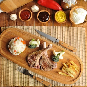 Könnte beinhalten: Ein Holzbrett mit Lammkoteletts, Reis, Gemüse und Pommes Frites. Enthält kleine Schalen mit Saucen, Knoblauch und einer roten Paprika. Ein Messer und eine Gabel sind ebenfalls vorhanden.