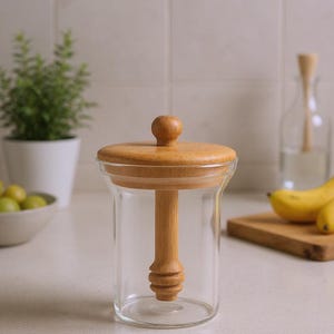 May include: A clear glass jar with a wooden lid and honey dipper. The lid has a small, round knob. The dipper is made of wood and has a decorative, grooved design. The jar is on a white countertop.