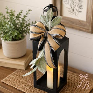 May include: A black metal lantern featuring a tan burlap bow with black and white gingham edges. The lantern is adorned with soft, light green lamb's ear leaves and contains a single white pillar candle emitting a warm, soft glow.