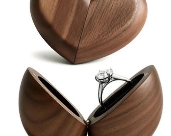 Op de afbeelding: Een donkerbruine, hartvormige houten ringdoos, opengeklapt om een zilveren ring met een heldere edelsteen te onthullen. De doos heeft een gladde afwerking en een zwart fluwelen interieur. De ring wordt in de geopende doos getoond.