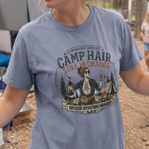 Op de afbeelding: Een stoffig blauw t-shirt met korte mouwen en een grafische print van een persoon die een lantaarn en een mok vasthoudt bij een tent en bomen. De tekst luidt CAMP HAIR STILL IN CHARGE en OUTDOOR OPERATIONS MANAGER in een cirkelvormig ontwerp.