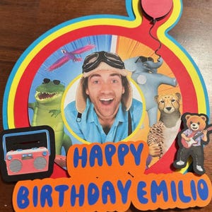 May include: A colorful birthday cake topper with the text "HAPPY BIRTHDAY EMILIO". The design includes a man wearing a pilot hat, cartoon animals, a radio, and a red balloon.