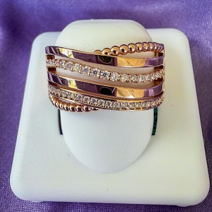 May include: Rose gold ring with multiple bands, some adorned with small, clear gemstones. The ring has a unique design with a beaded edge. The ring is displayed on a white stand against a purple background.