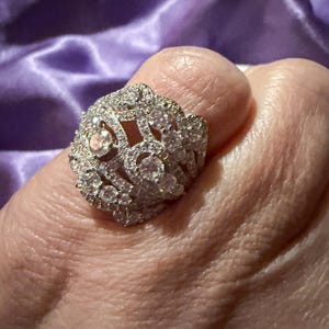 May include: A dazzling, ornate ring featuring a cluster of sparkling, clear gemstones. The ring has a detailed, openwork design and is set against a soft, purple background. The ring is a statement piece of jewelry.