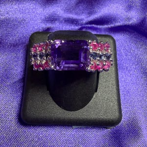 May include: A rectangular amethyst gemstone ring with pink and blue accent stones. The ring is set on a black display stand against a purple background. The amethyst is the primary focus, with the smaller stones adding color.
