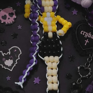 May include: A handmade keychain crafted from plastic pony beads in purple, yellow, black, and cream colors, arranged to form a stylized figure with a long purple ponytail, a yellow top, a black skirt, and black boots, attached to a metal clasp.