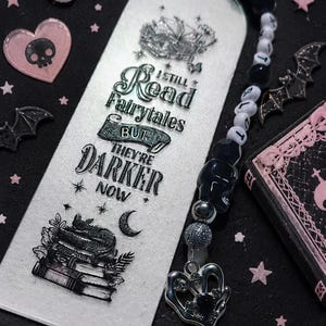 以下が含まれることがあります： 「I still read fairytales but they're darker now」のテキストが入ったゴシック調のブックマーク。 ビーズチャームとシルバーのハートペンダントが付いています。 画像にはピンクのハート、コウモリ、スカルが含まれています。