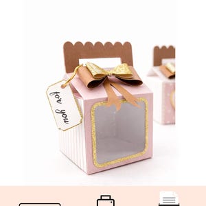 Può includere: Una scatola regalo rosa con una finestra trasparente e bordi glitterati dorati. La scatola ha un manico marrone e un fiocco dorato. Un'etichetta con la scritta "for you" è appesa alla scatola.