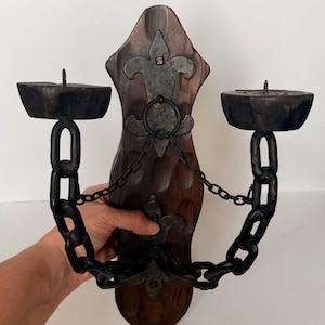 May include: A wall-mounted candle holder featuring a dark brown, carved wooden backplate with two metal fleur-de-lis accents. Two black metal chains extend from the centre to support circular candle platforms, each topped with a small metal spike.