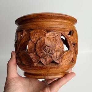 May include: A hand-carved wooden container with a floral design. The container is a warm brown colour and features intricate carvings of flowers and leaves. The design is cut out, creating a decorative effect.