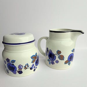 Peut inclure: Sucrier en céramique crème avec couvercle et pot à crème. Les deux pièces présentent un motif floral bleu et marron. Le sucrier a un bord et un couvercle bleu foncé. Le pot à crème a un bord noir et une anse.