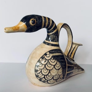May include: A ceramic jug shaped like a duck with a cream-coloured base and hand-painted black line details. The duck features a long neck, a curved handle on its back, and stylised feather patterns on its wings, finished with a glossy glaze.