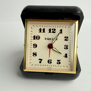 May include: A vintage travel clock with a cream-colored face, gold-tone frame, and black case. The clock face features black numerals, black hands, and the word "EQUITY". The clock is marked "CHINA".