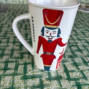 May include: A white ceramic Starbucks mug featuring a graphic of a nutcracker soldier in a red uniform with blue accents. The nutcracker has a tall hat, a mustache, and is winking. The word Starbucks is printed vertically in black along the handle.