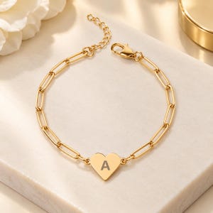 Peut inclure: Un bracelet à chaîne doré composé de maillons ovales allongés et d'une breloque centrale en forme de cœur. La breloque est gravée de la lettre majuscule A dans une police foncée. Le bracelet comprend un fermoir mousqueton et une chaînette.